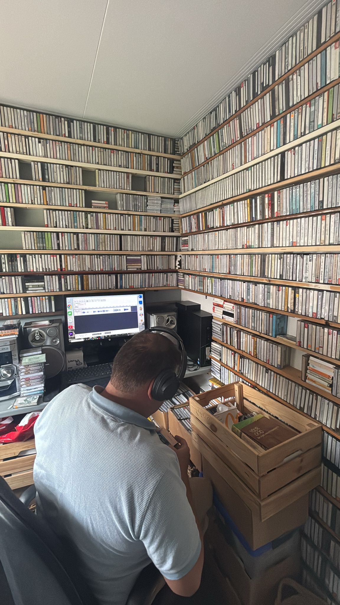 Willem met koptelefoon in zijn opnamehokje, omringd door wanden vol cassettebandjes